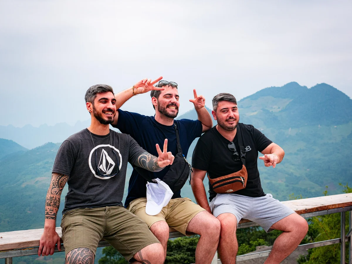 Three men sit on stools with mountains behind posing and smiling with hands in peace sign gestures