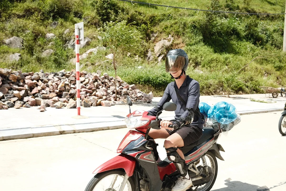 A man rides a red motorbike with a helmet on, a bag behind him, and there is sunshine