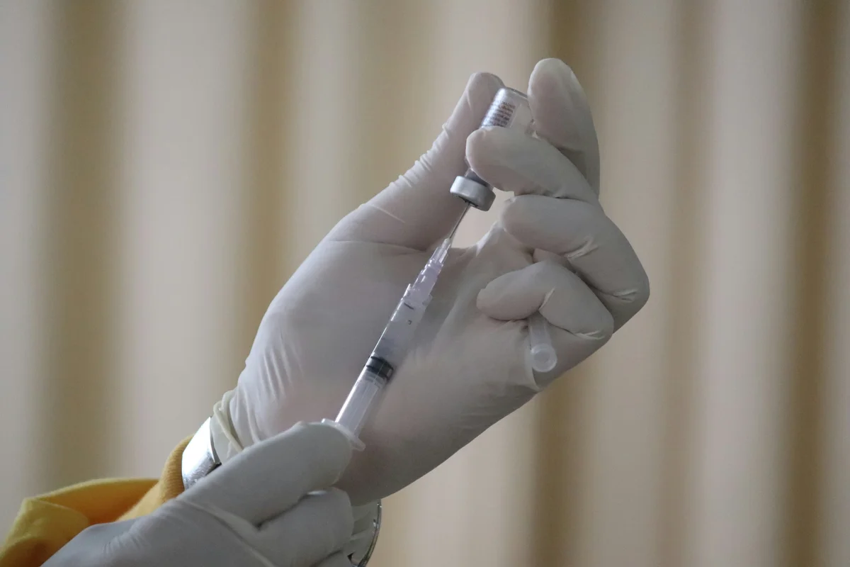 A hand holds a needle and vaccine bottle and extracts a small amount