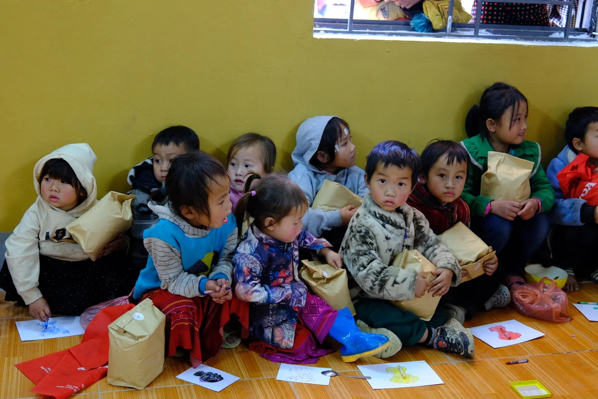 A group of children sit on the floor huddled together with coats on and papers on the floor around them