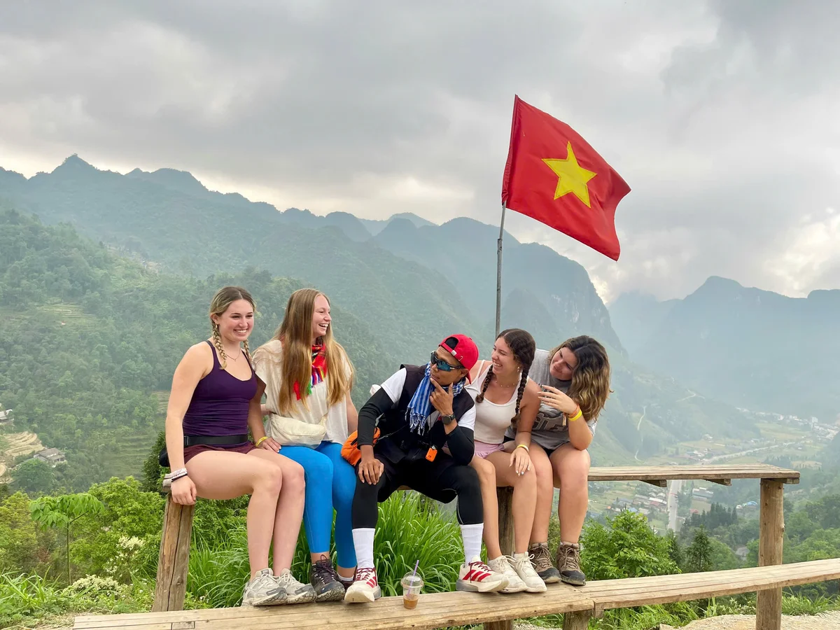 Group of girls smile and laugh with their Vietnamese easy rider at a viewpoint with a Vietnamese flag flying above and mountains behind