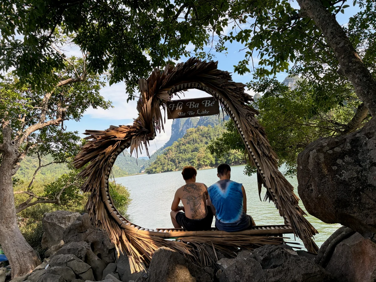 Two men sit in a heart shaped seat with trees surrounding them and a blue lake beyond