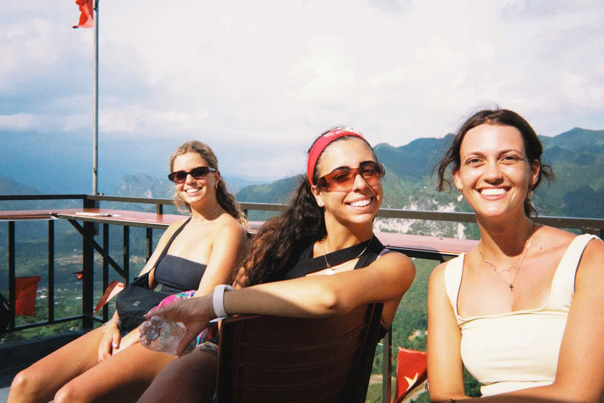Three women sit in the sunshine and smile at the camera with mountains and a railing behind them