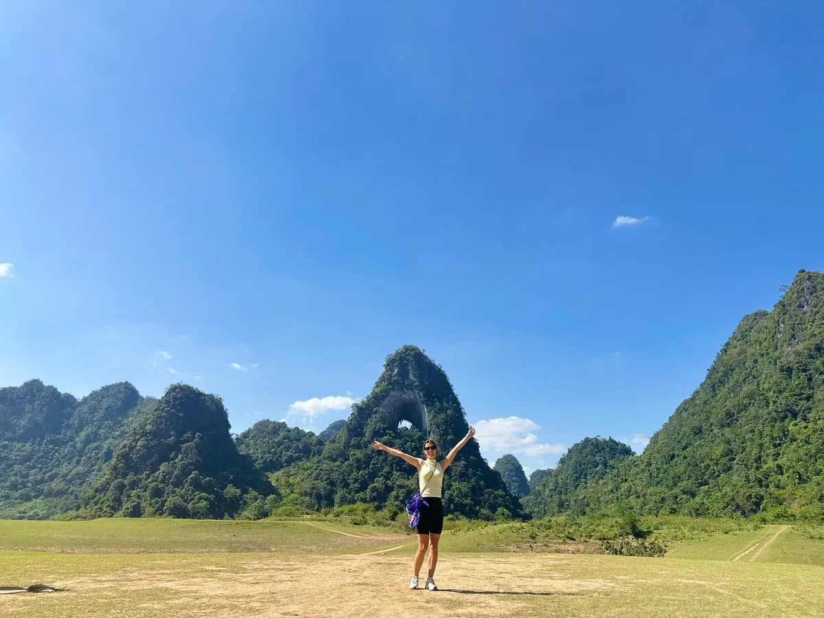A girl stands infront of a moutain with a hole in the middle with her arms spread wide