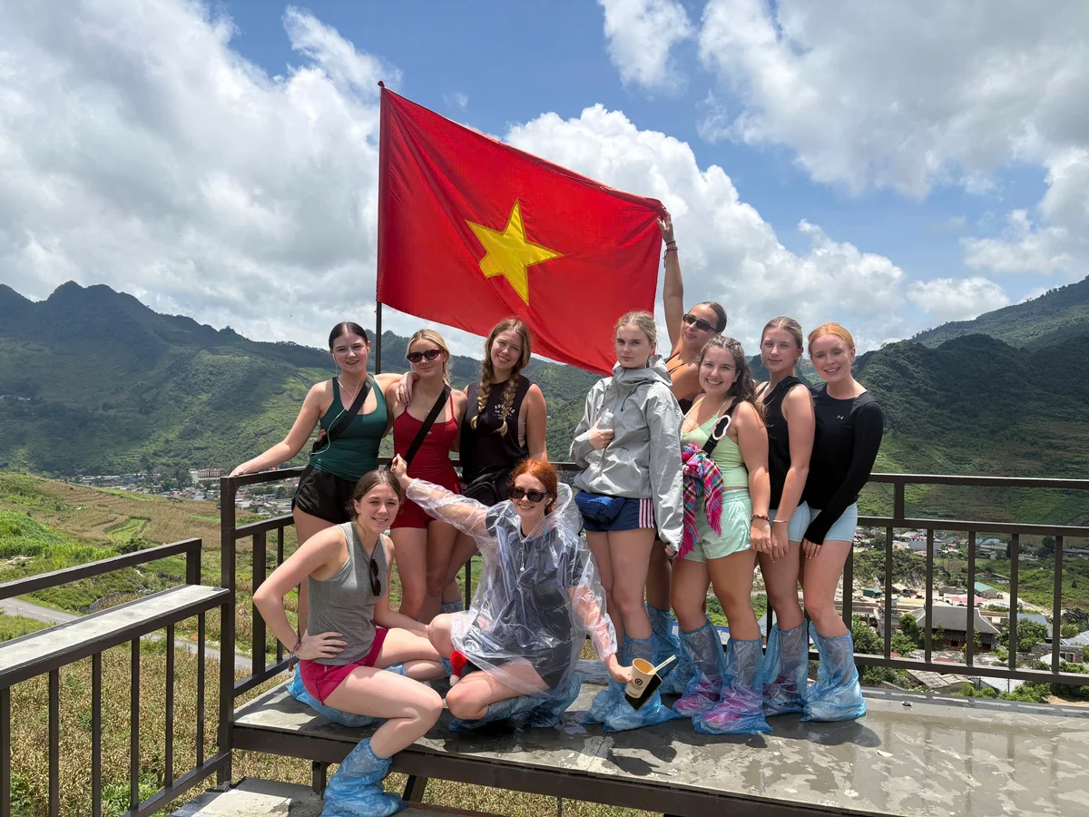 Sunshine, blue skies, and clouds, behind a group of people posing with a red flag with a yellow star. The people are dressed in ponchos, shorts, and light jackets.