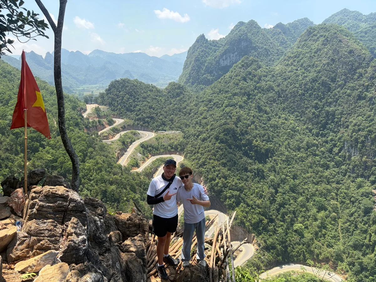 Two men stand on a cliff edge with a winding mountain road behind them and green mountains