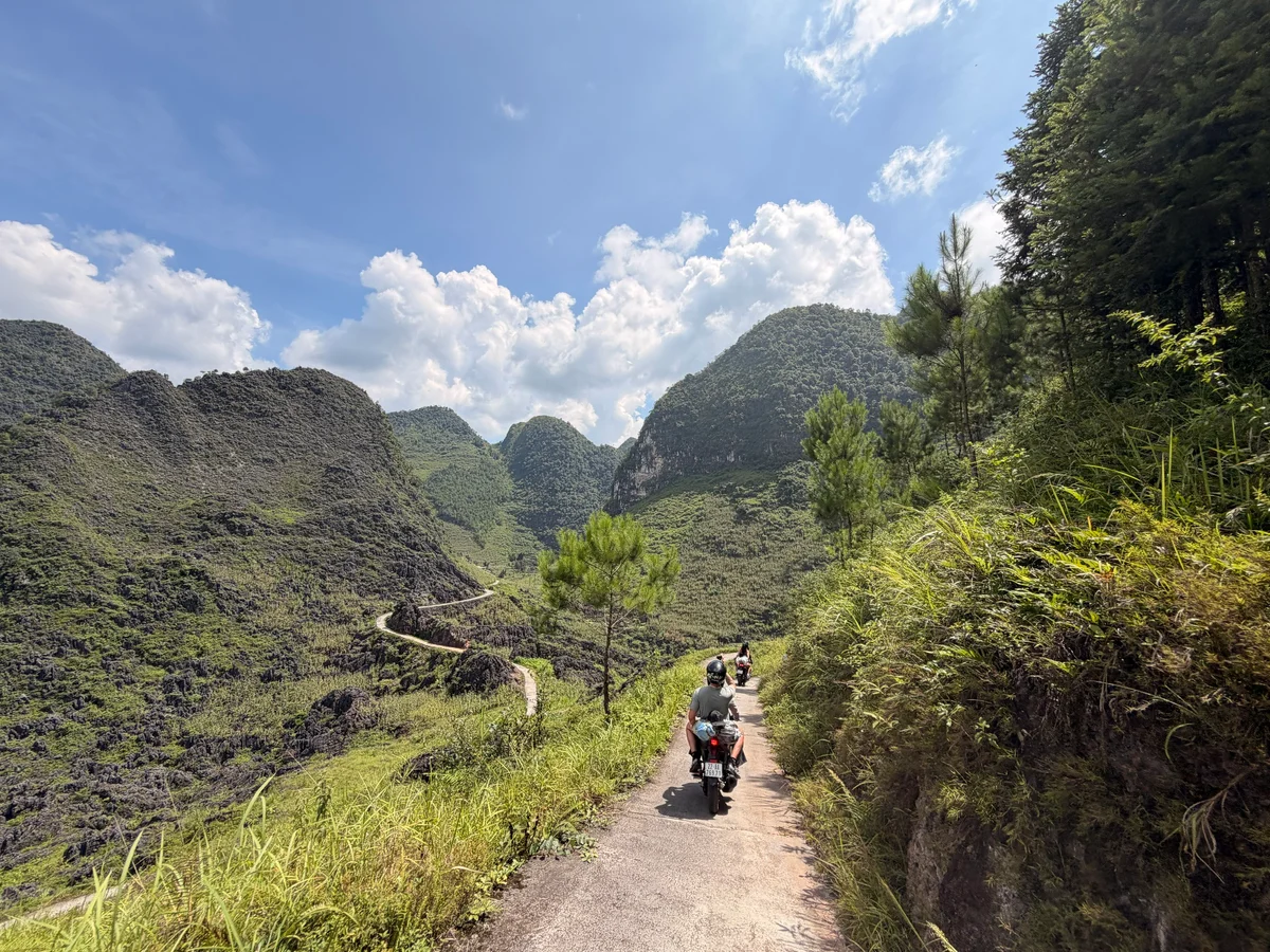 Sunshine, blue skies, and clouds as motorbikes ride through mountain scenery on a small road