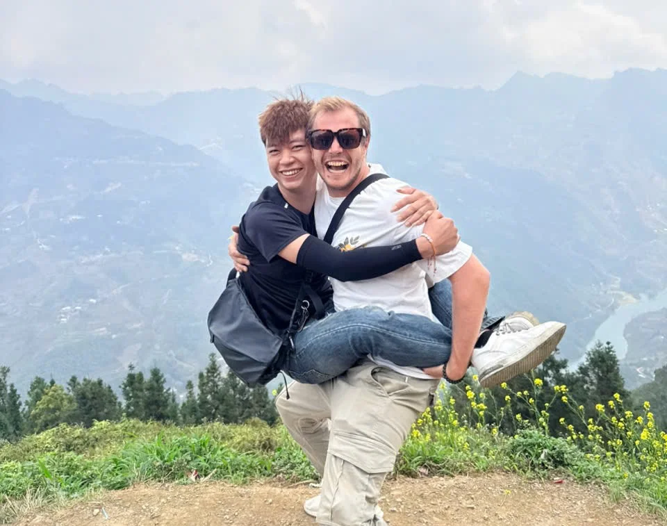 One man lifts another man up, both are smiling with mountains behind them and a blue river below