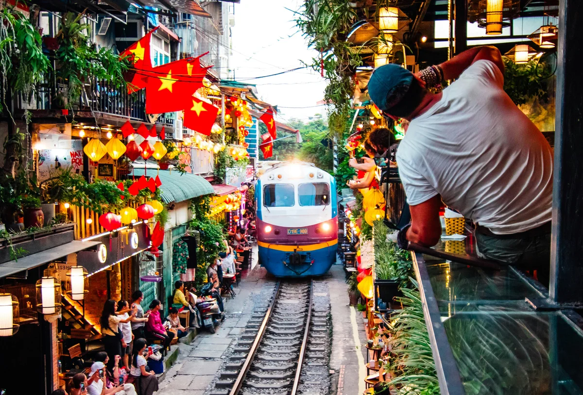 Train coming down the track with flags, lights, and lots of coffee shops and people either side