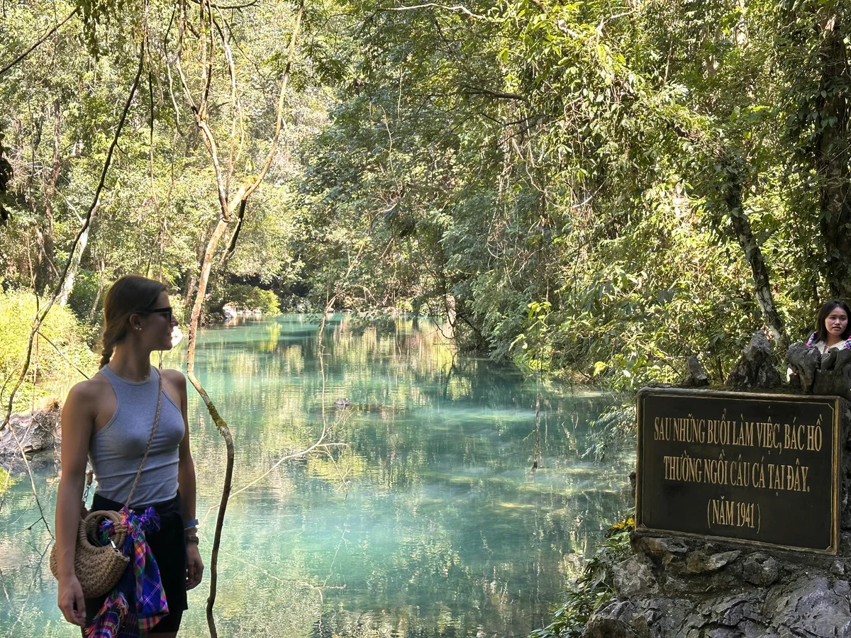 A girl stands next to a blue stream with trees all around