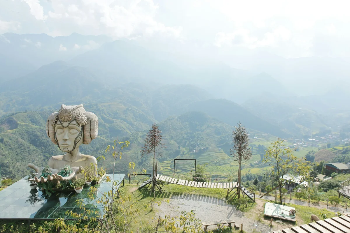 Landscape of mountains, rice fields, and a statue with outstretched arms, as well as benches and trees
