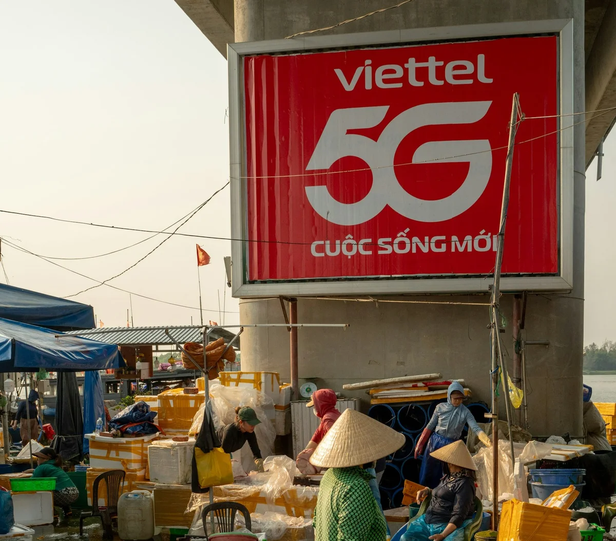 A stone pillar that has a Viettel 5G poster on it, there are people wearing conical hats standing at the base of the pillar