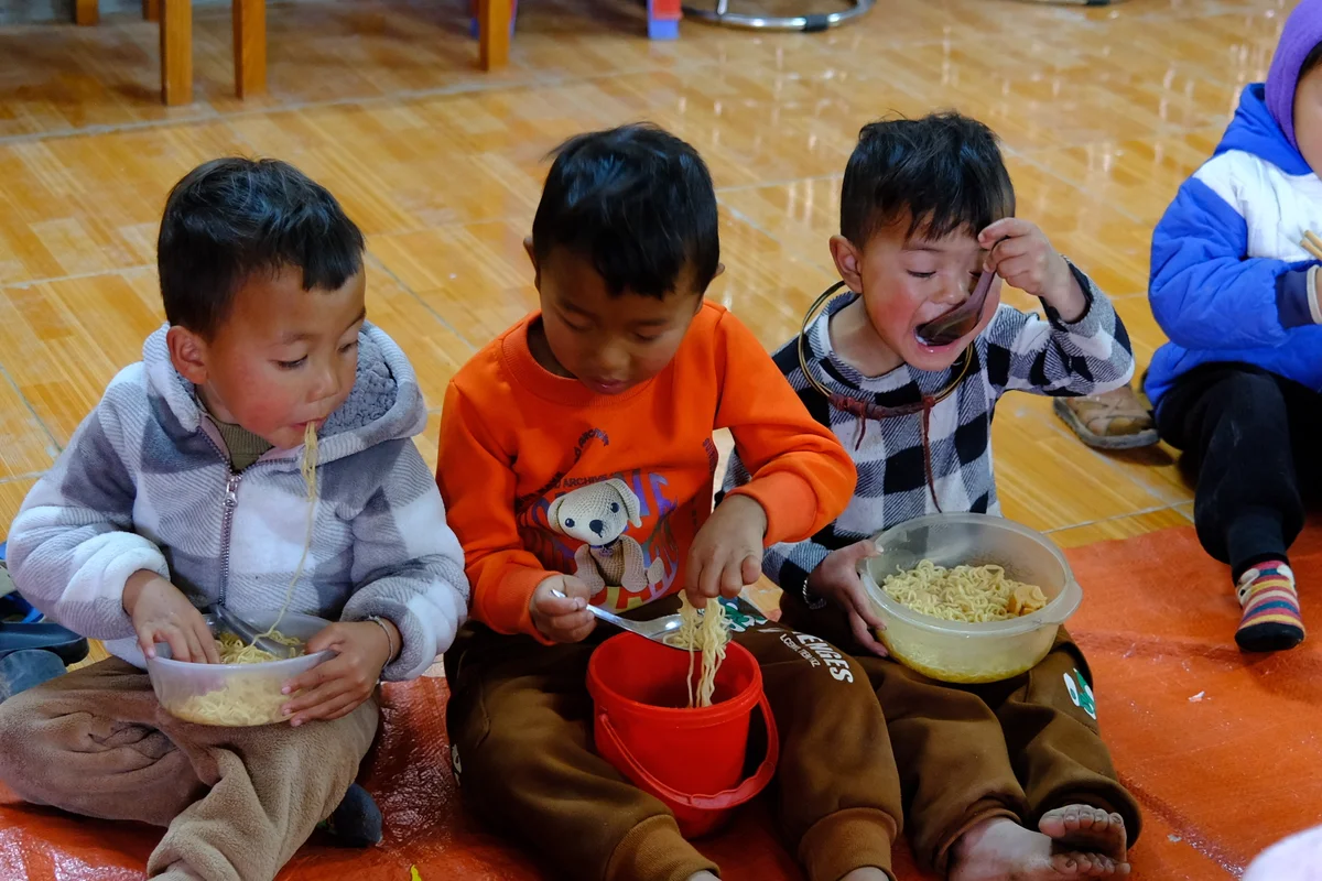 Three boys sit on the floor eating noodles